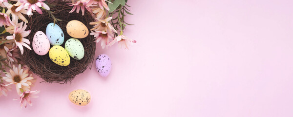 Obraz na płótnie Canvas Colorful easter eggs in nest and flowers on pink background with copy space. Flat lay. Banner.