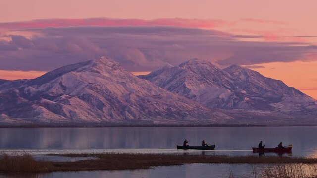 People in canoes floating on Utah Lake during colorful sunset looking towards Nebo mountain covered in snow.