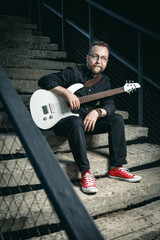 Obraz premium Portrait of a male guitar player with electric guitar wearing black clothing and red sneackers sitting on a stairs