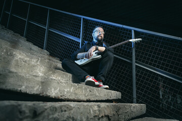 Portrait of a male guitar player with electric guitar wearing black clothing and red sneackers  sitting on a stairs