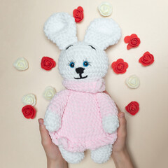 Handmade knitted toy. Knitted white rabbit in pink dress in hands on light background.