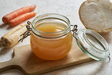 Bone broth in a glass jar on a white table, with vegetables