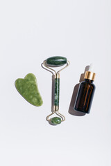 Jade face roller and glass bottle of beauty serum with shadows on on white background. Flat lay, top view