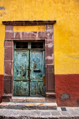 A very old faded turquoise wooden door with a stone frame and a yellow wall