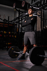 Fototapeta na wymiar male in sportive outfit having rest after hard exercises with barbell, handsome guy stands with folded arms looking at side. portrait of sportsman at modern gym