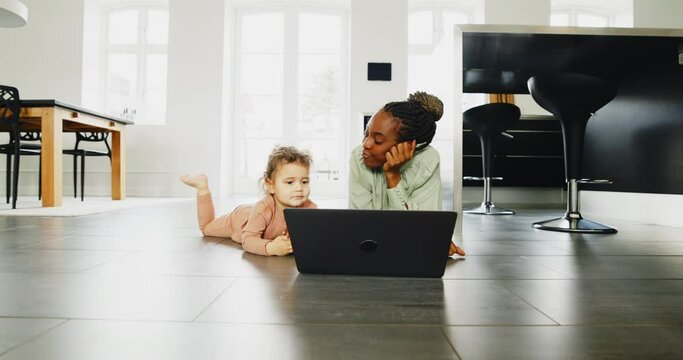 Smiling young African mom and her little daughter talking and watching something on a laptop while lying on their kitchen floor