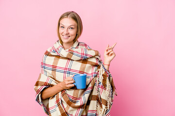 Obraz na płótnie Canvas Young russian woman wrapped in a blanket drinking coffee smiling cheerfully pointing with forefinger away.