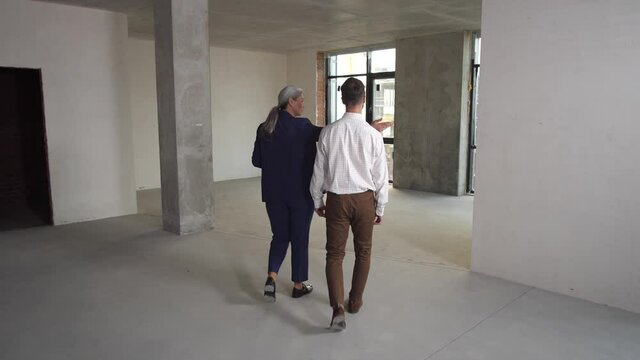 Rear view of woman realtor showing large apartment without finishing and interior partitions to client. Real estate agent and man buyer walking through empty space of apartment to see view from window