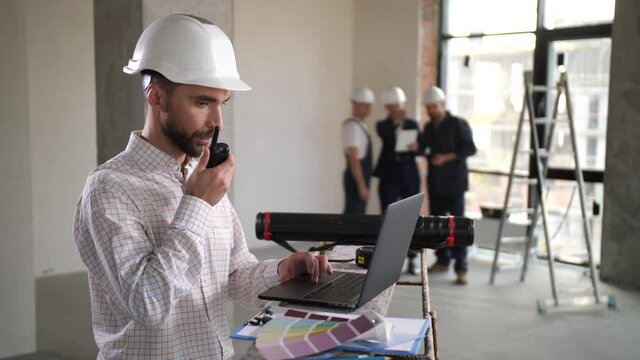 Serious architect working on laptop and using walkie-talkie while communicating with builders during apartment renovation. Designers and workers busy on renovation work in flat