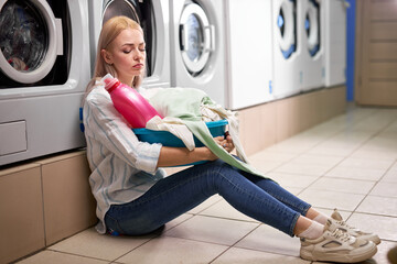 Bored woman holding dirty clothes and detergent pink bottle in basin, sits on the floor leaning on washing machine, sit depressed alone, in washing house