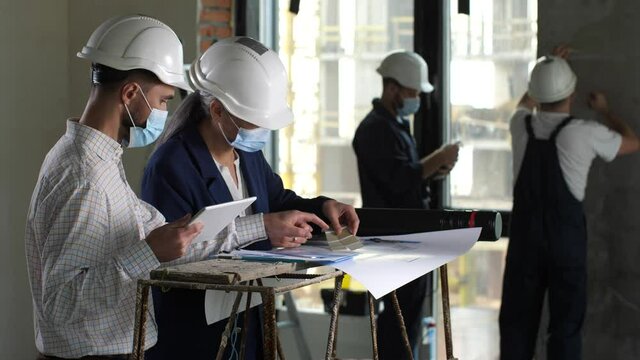 Professional architects in face masks and helmets discussing color scheme of apartment interior painting. Woman designer using color palette and consulting with man while choosing colors for interior