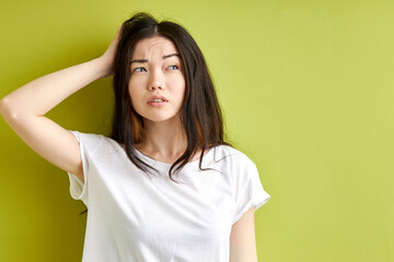 Fototapeta premium brunette woman try to remember something, stand thinking in contemplation touching hair and looking at side, female in casual t-shirt isolated over green studio background