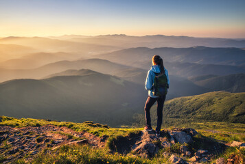 Girl on mountain peak with green grass looking at beautiful mountain valley in fog at sunset in summer. Landscape with sporty young woman, foggy hills, forest, sky. Travel and tourism. Hiking - obrazy, fototapety, plakaty