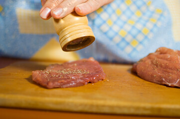 A woman peppers a slice of meat with a pepper mill. Cooking a meat chop using spices.