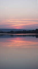 Blurry hazy image of a calm lake at sunset on a summer evening. Reflection in the water of delicate pastel colors of the sky. Artificial noise and graininess. Beauty of nature. Soft focus. Copy space