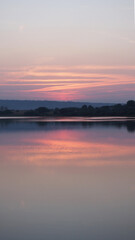 Fototapeta na wymiar Blurry hazy image of a calm lake at sunset on a summer evening. Reflection in the water of delicate pastel colors of the sky. Artificial noise and graininess. Beauty of nature. Soft focus. Copy space