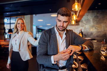 Unfaithful middle-aged man sitting in the bar of a fancy hotel and taking off his wedding ring. In the background is a woman who is seducing him.