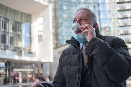 Man Drinking An Espresso Coffee While Sitting Outdoor In A City Square, Mask On His Chin, Covid Coronavirus Concept