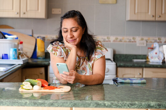 Hispanic Mom Searching Recipes On Her Phone-woman Preparing Healthy And Organic Salad While Checking Her Cell Phone-bored Housewife While She Looks At Her Phone