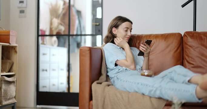 Young and cheerful woman reading on phone at home, sitting on the comfortable sofa in a cozy and stylish living room