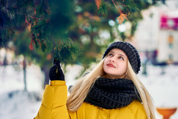 A pretty blonde with long hair and a yellow down jacket touches the paw of a huge fir tree.