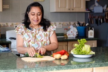 Smiling middle aged hispanic woman making fresh salad in her kitchen - lady in cooking healthy food...