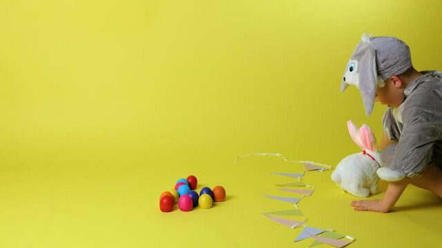 little cheerful boy in bunny grey costume with bunny ears playing with toy rabbit, colorful eggs on trendy ultimate yellow background, slow motion, Happy And Fun Easter Day concept celebrate
