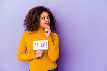 Young African American woman holding a Help placard isolated on purple background looking sideways with doubtful and skeptical expression.