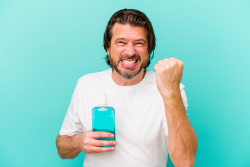 Middle age dutch man sitting holding a mouthwash isolated on blue background showing fist to camera, aggressive facial expression.