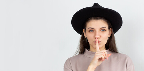 Woman ready to tell secret of beauty. Portrait of charming flirty caucasian female making shush or shh gesture with index finger over mouth, looking sensually and confident at camera over white wall