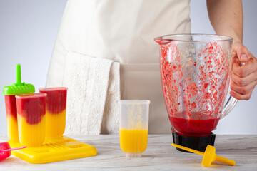 A mother is preparing homemade all natural fruit popsicles. She is filling bpa free plastic molds with layers of pureed mango and berry medley before freezing them. She holds mixer bowl with puree.