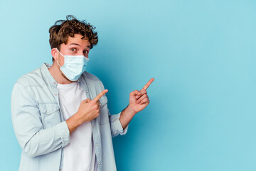 Young caucasian man wearing an antiviral mask isolated on blue background pointing with forefingers to a copy space, expressing excitement and desire.