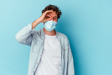 Young caucasian man wearing an antiviral mask isolated on blue background excited keeping ok gesture on eye.