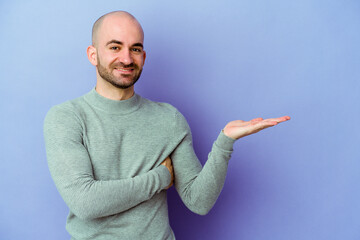 Young caucasian bald man isolated on purple background showing a copy space on a palm and holding...