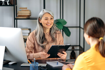 A Mature gray-haired asian woman, employer, examines the resume of a female job seeker, sitting at...