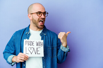 Young caucasian bald man holding a made with love placard isolated on purple background points with thumb finger away, laughing and carefree.