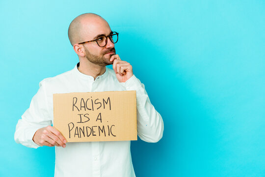 Young caucasian bald man holding a racism is a pandemic isolated on white background looking sideways with doubtful and skeptical expression.