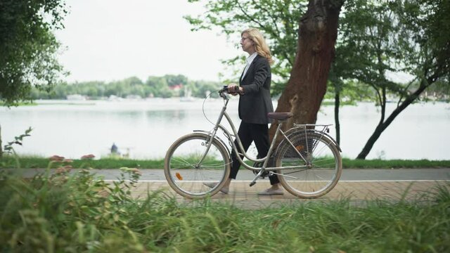 Side view of happy relaxed European businesswoman walking with bicycle along lake shore in urban park. Wide shot of confident slim Caucasian blond middle aged woman enjoying strolling outdoors.