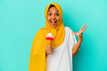 Young muslim woman eating an ice cream isolated on blue background receiving a pleasant surprise, excited and raising hands.