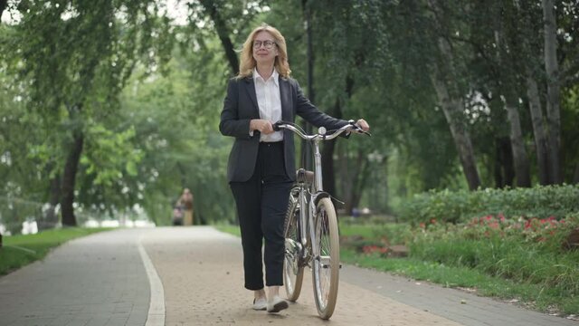 Wide shot portrait of smiling successful Caucasian businesswoman walking with bike on summer park alley. Happy relaxed middle aged woman in eyeglasses and suit strolling on break outdoors.