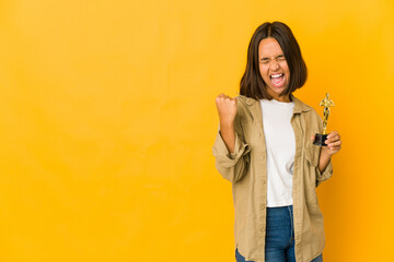 Young hispanic woman holding an award statuette raising fist after a victory, winner concept.