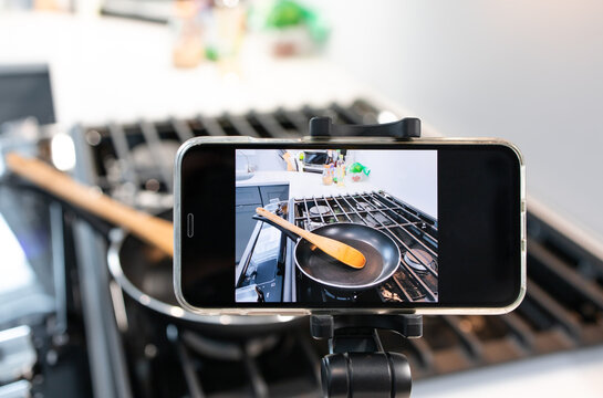 Mobile phone on tripod with video record screen of kitchen utensils on gas range and blurred seasoning of food in the kitchen for cooking meal at home, preparation for blogger live at online media.