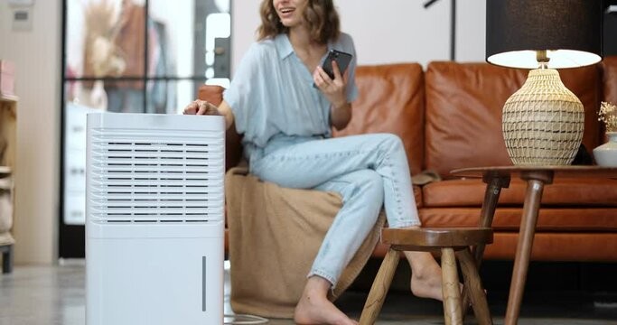 Woman switching on portable air conditioner while sitting on a couch at home. Enjoying fresh and clean air at home. Home appliances for cleaning and air conditioning
