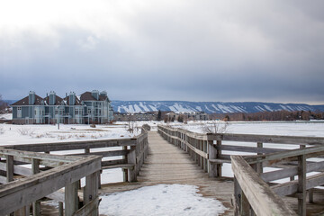 A wooden bridge in Lighthouse Point, Collingwood, crosses a portion of Georgian Bay while overlooking the Blue Mountain Ski hills from a distance, with the frozen water beside it