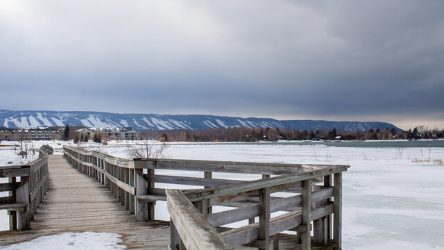 A Wooden Bridge In Lighthouse Point, Collingwood, Crosses A Portion Of Georgian Bay While Overlooking The Blue Mountain Ski Hills From A Distance, With The Frozen Water Beside It