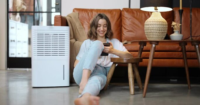 Woman controlling with smartphone portable air conditioner while sitting on the floor at home. Enjoying fresh and clean air at home. Smart home appliances for air cleaning and conditioning