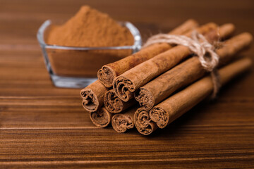 Aromatic cinnamon sticks and glass bowl with powder on wooden table, closeup