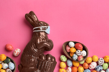 Chocolate bunny with protective mask and eggs on pink background, flat lay. Easter holiday during COVID-19 quarantine