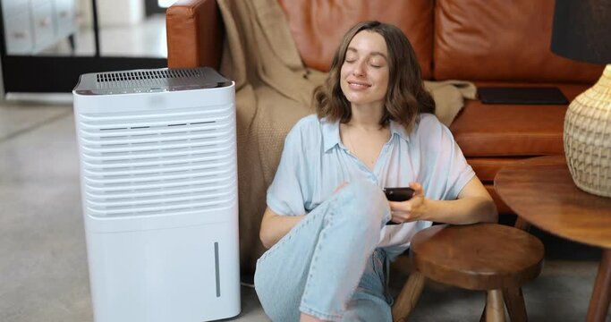 Woman controlling with smartphone portable air conditioner while sitting on the floor at home. Enjoying fresh and clean air at home. Smart home appliances for air cleaning and conditioning