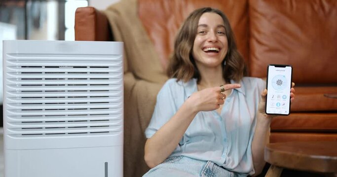 Happy Woman Controlling With Smartphone Portable Air Conditioner, Showing Device With A Running Program. Enjoying Fresh And Clean Air At Home. Smart Home Appliances For Air Cleaning And Conditioning